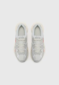 Lichtgrijze sneakers met een mesh- en suède textuur, afgeronde vorm, contrasterende witte panelen en een rode accent aan de zijkant. Voorzien van platte veters.