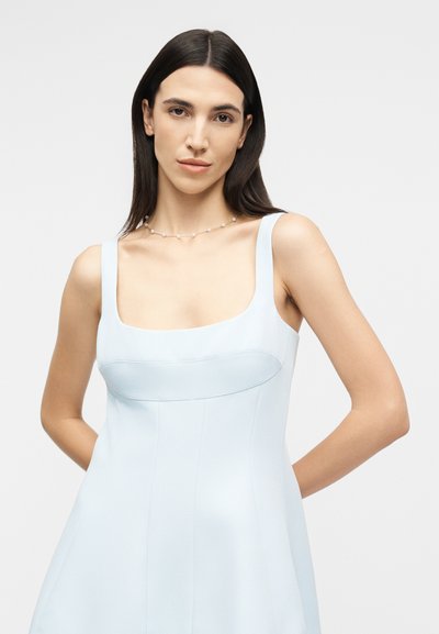 Femeie cu păr lung și întunecat, purtând o rochie albastru deschis fără mâneci și un colier delicat de perle, stând în fața unui fundal alb simplu.