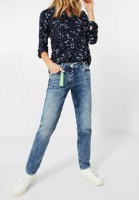 Námořnická květinová košile s dlouhým rukávem, se vzory v bílé a modré barvě, spárovaná se světle modrými džíny a bílými teniskami, na které je zelený štítek na pasu.