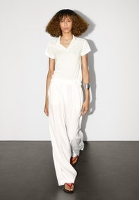 Bílá polokošile doplněná volnými bílými kalhotami. Výrazný stříbrný náramek na ruce. Obuv zahrnuje růžové a červené žabky.