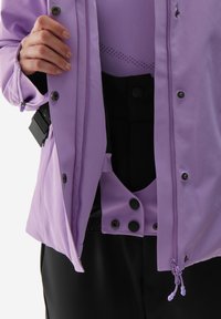 Fialová bunda vyrobená z hladkého materiálu, s černými knoflíky a kapsou na zip. Pod ní je přiléhavý fialový top s síťovaným vzorem.