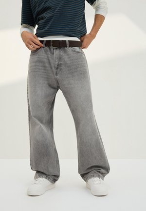 Personne portant un jean gris délavé à jambes larges, des baskets blanches, une ceinture marron foncé et une chemise rayée bleu foncé avec des poignets blancs.