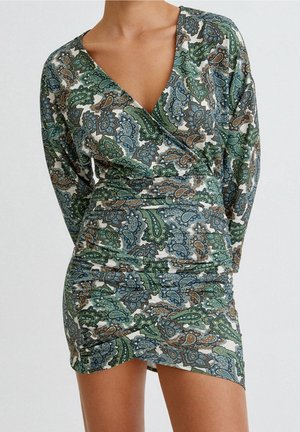 Femme portant une robe cache-cœur à manches longues avec des motifs cachemire verts, bleus et bruns sur un fond clair.