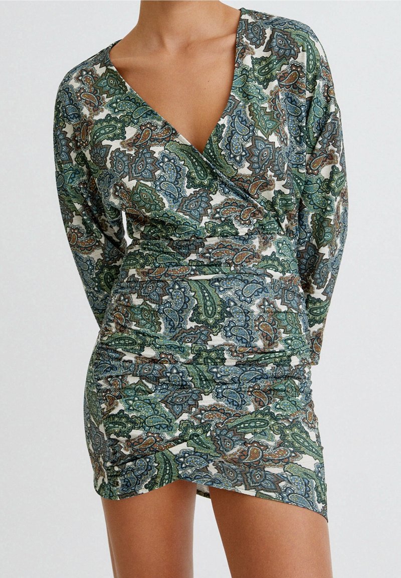 Femme portant une robe cache-cœur à manches longues avec des motifs cachemire verts, bleus et bruns sur un fond clair.