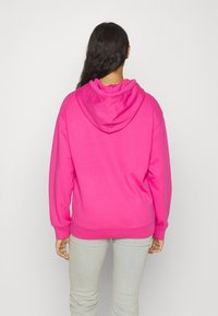Felpa rosa con cappuccio, maniche lunghe e polsini a costine. Colore uniforme senza motivi. Abbinata a pantaloni aderenti grigio chiaro.
