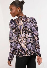 Caurspīdīga melna blouse ar purpura un zelta ziedu rakstu, sagriezta kakla izgriezumā un pūkainām piedurknēm. Melni pieguļoši bikses noslēdz izskatu.