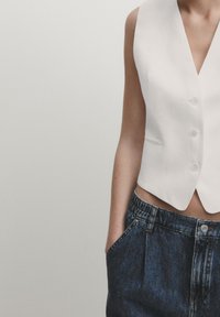 Biała bezrękawnikowa kamizelka z głębokim V-kształtnym dekoltem, ozdobiona czterema guzikami i dwoma bocznymi kieszeniami, w zestawieniu z wysokim stanem szortów dżinsowych.