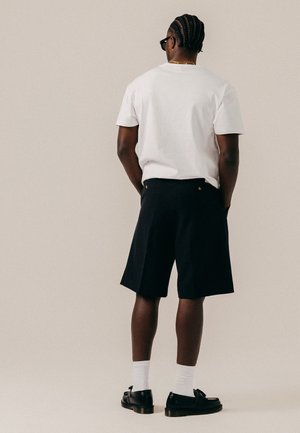 Homme aux cheveux tressés portant des lunettes de soleil noires, un t-shirt blanc, un short noir, des chaussettes blanches et des mocassins noirs, debout les mains dans les poches.
