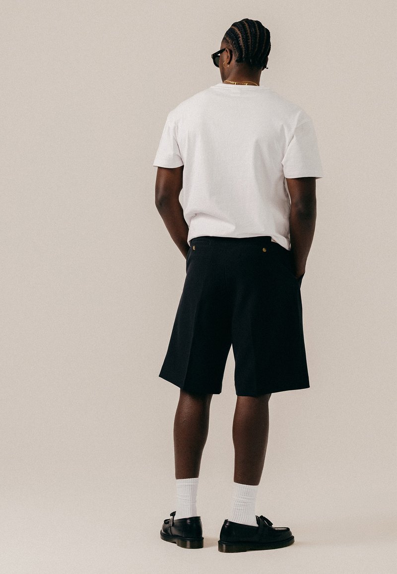 Homme aux cheveux tressés portant des lunettes de soleil noires, un t-shirt blanc, un short noir, des chaussettes blanches et des mocassins noirs, debout les mains dans les poches.