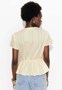 Blouse peplum jaune clair avec des manches courtes structurées, en tissu texturé avec des motifs subtils, taille cintrée et ourlet évasé.