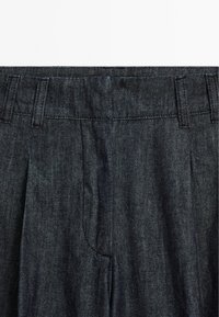 Donkerdenim broek met een platte voorkant, dubbele plooien en riemlussen. De stof heeft een iets ruwe textuur met subtiele variaties.