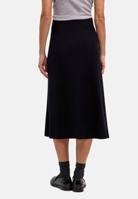 Gonna midi nera con una texture liscia, vita aderente e design svasato. Presenta una chiusura con zip sul retro. Indossata con calze scure e scarpe lucide.