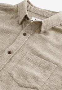 Chemise beige texturée à boutons avec col, deux boutons marron et une poche poitrine sur fond blanc.