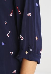 Blouse marine à manches retroussables, ornée de motifs brodés colorés de étoiles, cœurs, guitares et lèvres sur un tissu doux.