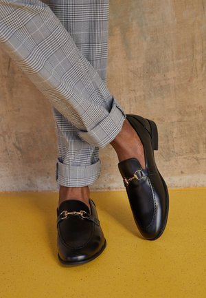 Mocassins en cuir noir avec un détail chaîne dorée, associés à un pantalon gris à carreaux. Détails visibles comprenant une texture lisse et une forme structurée.