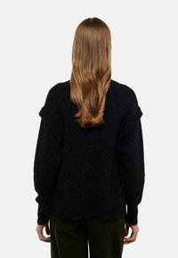 Personne avec de longs cheveux ondulés châtain clair portant un pull noir et un pantalon sombre, se tenant de dos contre un fond clair uni.