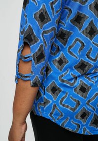 Zila rakstota blūze ar ģeometriskiem pašiem melnā un baltā krāsā, ar izgrieztām piedurknēm un gludu tekstūru.