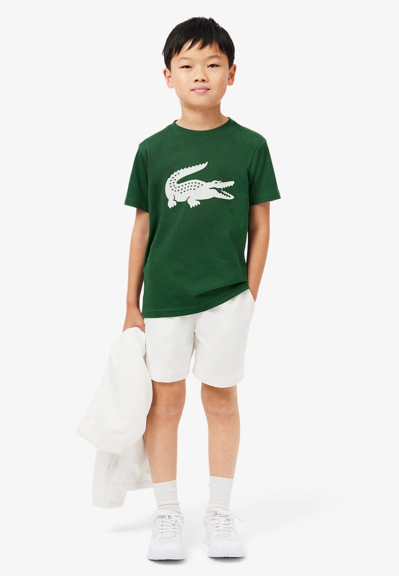 T-shirt en coton vert avec un graphique de crocodile blanc. Associé à un short blanc et des baskets blanches. Tient une veste blanche.