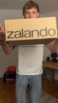 Καφέ κουτί από χαρτόνι με την ένδειξη "zalando" τυπωμένη σε μεγάλα μαύρα γράμματα, κρατημένο από ένα άτομο με λευκό πουκάμισο και μπλε τζιν.