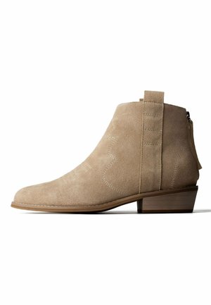 Bottine beige en daim avec talon bas empilé, languette latérale, coutures décoratives et fermeture éclair au dos.