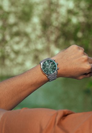 Polso con orologio a cinturino metallico argento che mostra quadrante verde con più quadranti secondari, su sfondo naturale sfocato.