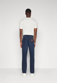 Σκούρα μπλε τζιν με ίσια γραμμή, με καφέ δερμάτινη πινακίδα στο πίσω μέρος, σε συνδυασμό με ένα ανοιχτόχρωμο T-shirt και λευκά αθλητικά παπούτσια.