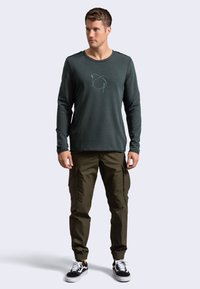 Camicia a maniche lunghe di un verde scuro con un sottile grafica astratta, abbinata a pantaloni cargo color oliva. Modello che indossa sneakers nere.
