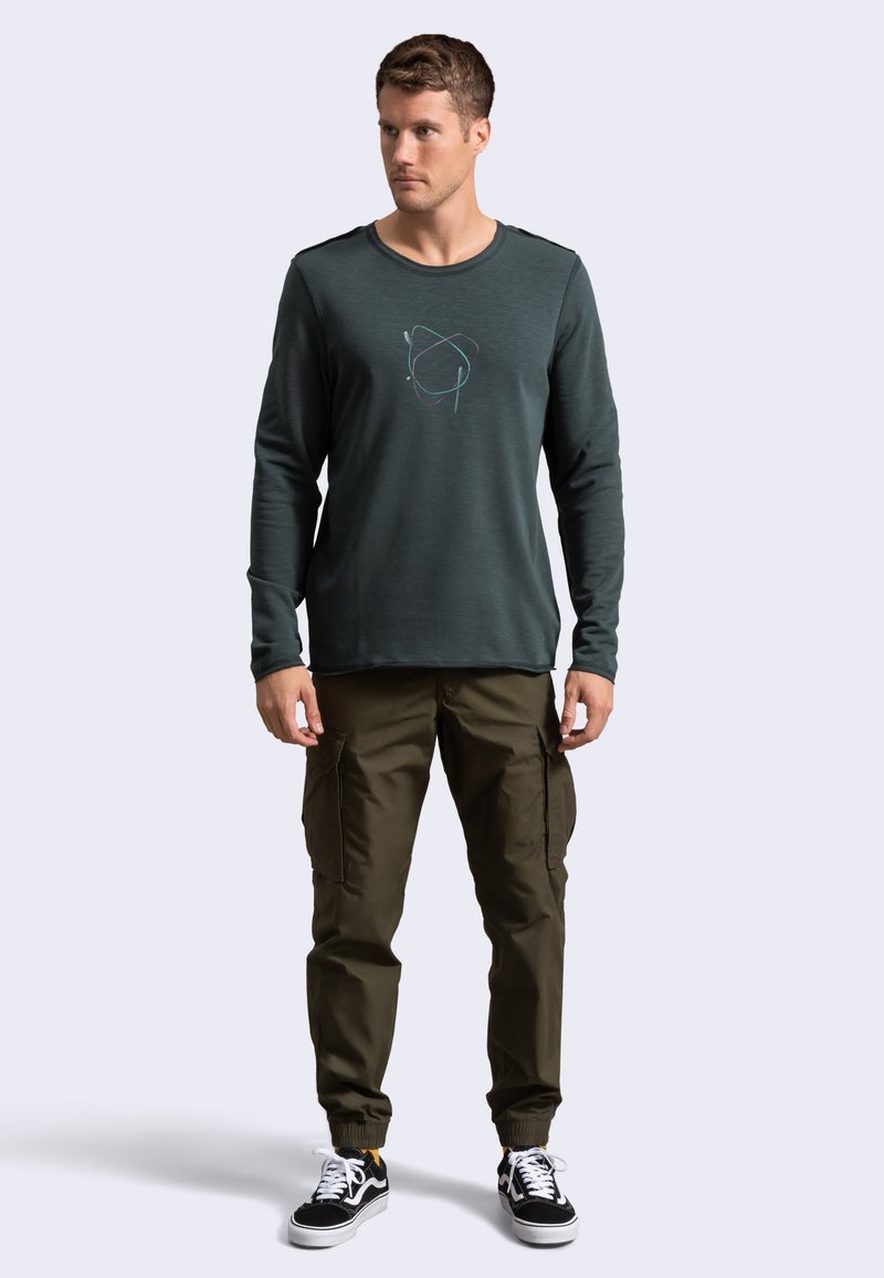 Camicia a maniche lunghe di un verde scuro con un sottile grafica astratta, abbinata a pantaloni cargo color oliva. Modello che indossa sneakers nere.