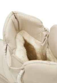 Intérieur de botte beige avec une doublure en fleece douce et claire, bords rembourrés et matériau extérieur lisse. Détail de couture visible.