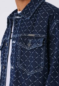 Chaqueta de mezclilla azul oscura con un patrón geométrico en hilo blanco, con un cuello estándar y un bolsillo en el pecho con botones. Detalles de hardware en plata visibles.
