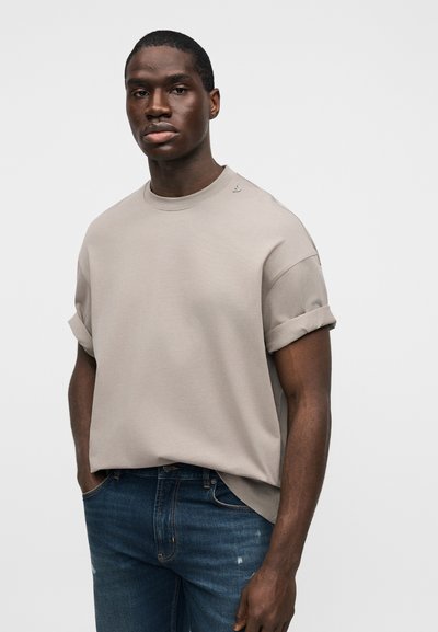 Νεαρός άνδρας που φοράει μπεζ φαρδιά κοντομάνικη πουκάμισο με γυρισμένα μανίκια και σκούρο μπλε τζιν, όρθιος με το ένα χέρι στην τσέπη.