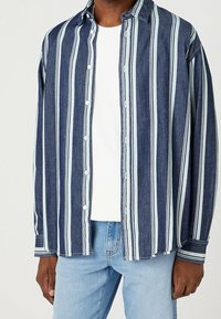 Person med en blå och vit vertikalt randig skjorta över en vit t-shirt och ljusblå jeans, stående mot en enfärgad bakgrund.