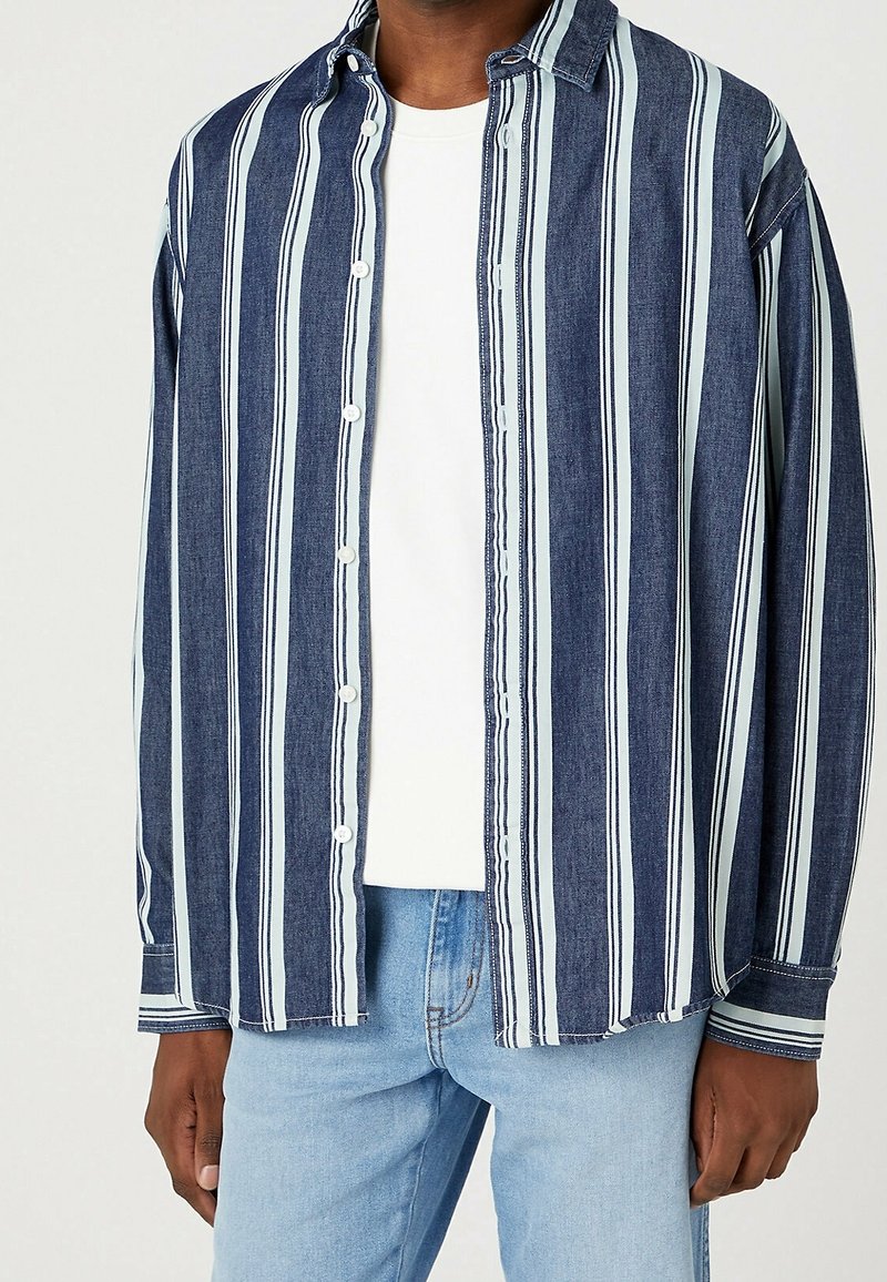 Person med en blå och vit vertikalt randig skjorta över en vit t-shirt och ljusblå jeans, stående mot en enfärgad bakgrund.