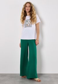 Witte T-shirt met een bloemig ontwerp in het midden van de borst, in combinatie met groene wijde broekspijpen van een gestructureerde stof. De schoenen zijn lichtgekleurde sandalen.