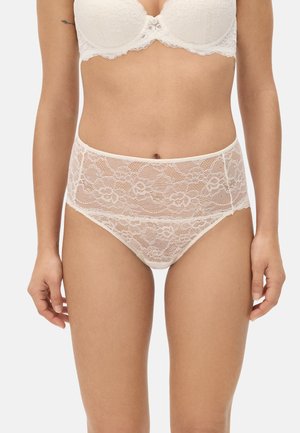 Mujer con ropa interior blanca de encaje de talle alto y sujetador a juego, de pie con los brazos relajados a los lados contra un fondo liso.