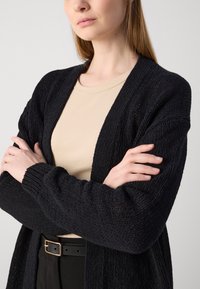 Černý pletený kardigan s volným střihem, který má texturovaný vzor a otevřený přední díl. Nosič má na sobě světle béžové tričko s dlouhými rukávy.