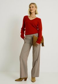 Un pull rouge à col en V associé à un pantalon large à motifs vichy beige. L'accessoire comprend une pochette en daim marron avec des grelots. Des bottes à talons chunky.