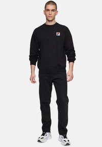 Felpa nera con colletto rotondo e maniche lunghe, caratterizzata da un piccolo logo rosso, bianco e blu. Abbinata a jeans neri e sneaker bianche.