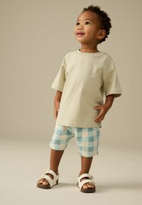 T-shirt beige clair à manches courtes, short à carreaux bleus et blancs, et sandales blanches avec une boucle. Tenue pour enfants simple et confortable.