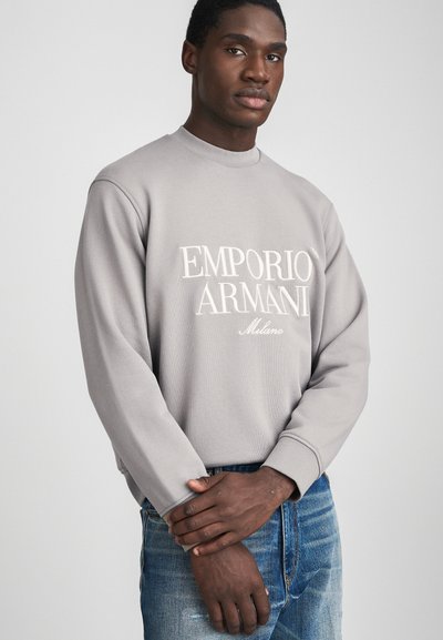 Felpa grigia con maniche lunghe e colletto a coste. Presenta il logo bianco "EMPORIO ARMANI Milano" sulla parte anteriore. Indossata con jeans blu.