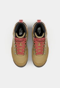 Bottes de randonnée beiges avec des lacets rouges, œillets renforcés et une semelle en caoutchouc texturée. L'intérieur présente une doublure noire et un logo.