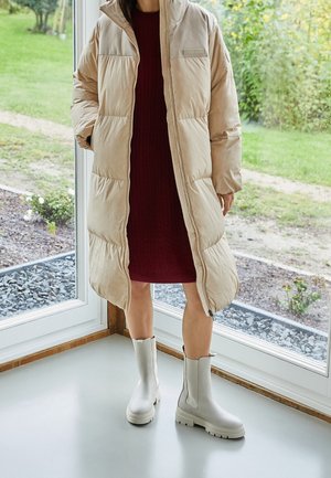 Puffersjakke i lyse beige med lynlås foran, med høj krave og foret. Båret over en rød strikkjole og parret med lysegrå ankelstøvler.