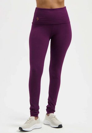 Lila Leggings mit einem breiten, umgeschlagenen Bund und einer glatten Textur. Verfügt über ein dezentes Marken-Detail in Orange. Kombiniert mit hellen Sneakers.