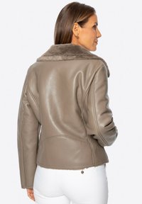 Taupe leren jack met een zacht bontkraag, op maat gesneden, en zichtbare stiksels, gedragen met witte broek. Glad textuur.