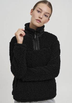 Schwarzer Fleece-Pullover mit hohem Kragen, Reißverschluss vorne und strukturiertem Oberflächenmaterial. Verfügt über ein kontrastierendes glattes schwarzes Paneel auf der Brust.