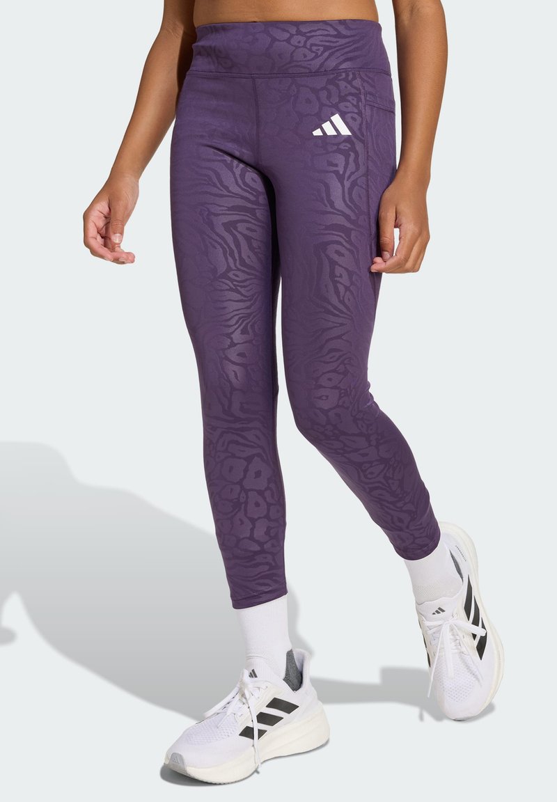 Leggings violets avec un imprimé animal texturé, dotés d'une taille haute et du logo Adidas en blanc, associés à des chaussures de sport blanches.