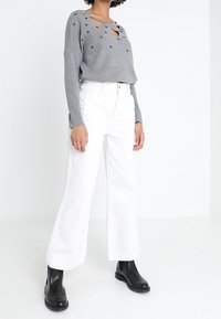 Grijze lange mouwen gebreid top met uitgesneden halslijn en versieringen, gecombineerd met hoge taille witte wijde broek en zwarte enkellaarsjes.