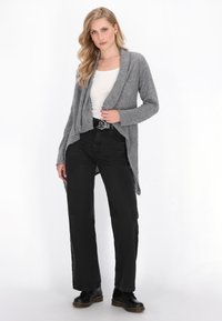 Una donna è in piedi indossando un cardigan grigio aperto sul davanti, una maglietta bianca, jeans neri a vita alta, una cintura nera e scarpe nere con lacci su uno sfondo bianco.