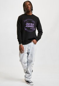 Svart hettegenser med lilla grafisk logo i midten, kombinert med lys blå baggy jeans og svarte joggesko. Modellen står mot en enkel bakgrunn.