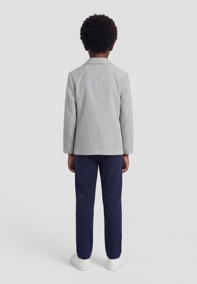 Enfant debout, dos tourné, portant un blazer gris à fines rayures, un pantalon bleu marine et des baskets blanches sur un fond uni.